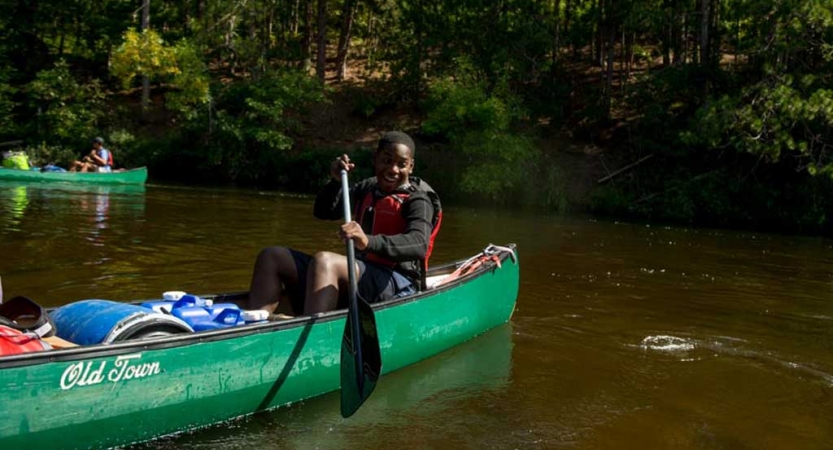 A person wearing a life jacket paddles a canoe on calm water near a tree-lined shore.